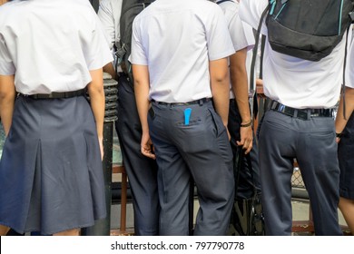 Back view of Thai university or vocation student in uniform, navy blue trouser or skirt and white shirt, backpack. One man has blue bomb in pocket. Education concept.