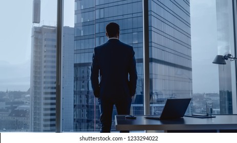 Back View of the Thoughtful Businessman wearing a Suit Standing in His Office, Hands in Pockets Looking out of the Window. Big City Business District Panoramic Window View. Dark Rainy Weather. - Powered by Shutterstock