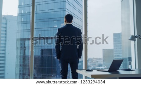 Back View of the Thoughtful Businessman wearing a Suit Standing in His Office, Hands in Pockets and Contemplating Next Big Business Deal, Looking out of the Window. Big City Business District View