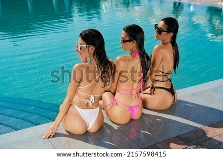 Back view of three seductive girl sitting near swimming pool, sunbathing. Pretty slim young women looking down, relaxing, having vacation. Concept of summertime and youth.