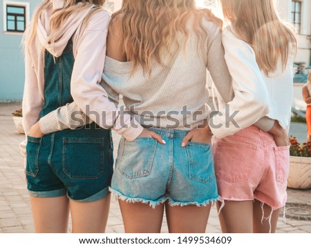 Back view of three young female hipster friends.Girls dressed in summer casual clothes.Women standing outdoors.They put their hands in shorts in back pockets.Posing at sunset