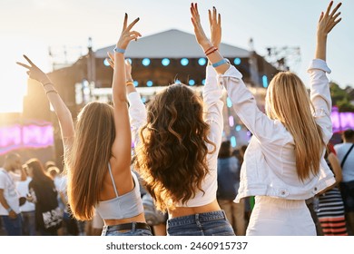 Back view of three young women at beach music festival, arms raised, dancing, enjoying summer sunset party vibe. Friends celebrate, fun outdoor event, casual fashion, stage lights in background. - Powered by Shutterstock