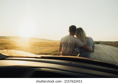 Back View Through Front Car Window on Young Couple Hugging Near the Hood, Woman and Man Enjoying Road Trip at Sunset - Powered by Shutterstock