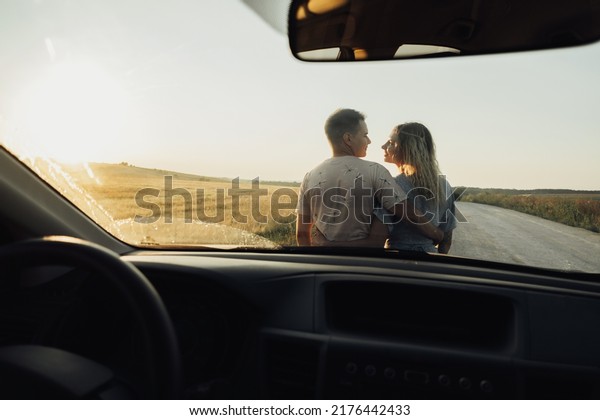 Back\
View Through Front Car Window on Young Couple Hugging Near the\
Hood, Woman and Man Enjoying Road Trip at\
Sunset