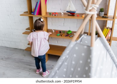 Back View Of Toddler Girl With Down Syndrome Reaching Toys On Wooden Shelf 