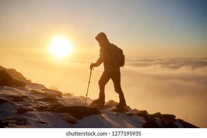 Back view of tourist hiker with backpack and hiking sticks hiking on rocky mountain peak on copy space background of beautiful foggy valley filled with white puffy clouds at dawn at winter.