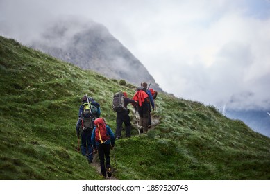 Back view of tourists with backpacks using trekking poles while climbing the grassy hill. Group of active people walking on path and heading to foggy mountain. Concept of hiking and backpacking.