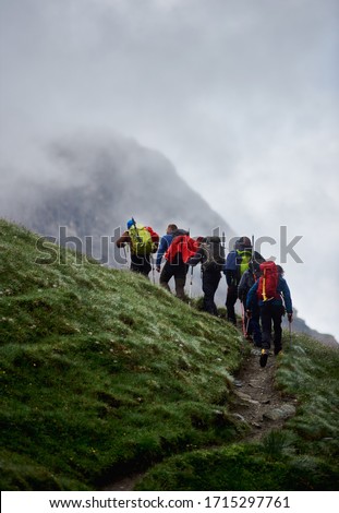 Back view of tourists with backpacks while climbing the grassy hill with foggy mountain on background. Hikers team with trekking sticks waking on hillside path. Concept of hiking and backpacking.