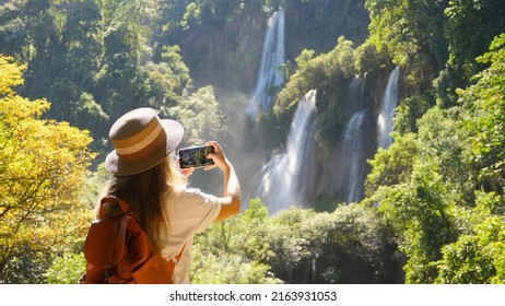 Back View Of Travel Blogger Woman On Trip Or Adventure On Tee Lor Su Waterfall In Thailand. Young Female Traveler Make Photos On Smart Phone For Social Media. Generation Z New Concept Travels