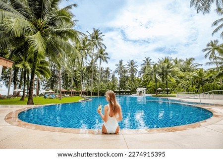 Back view of travel woman influencer with tropical cocktail sitting in swimming pool with palm trees and blue sky. Female traveler relax at luxury beach resort. Vacations and summer concept.
