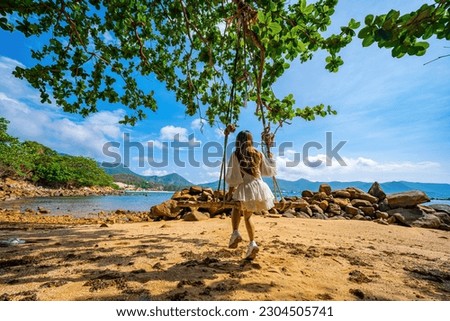 Back view of travel woman sitting on swing on the beach with palm trees and indian almond tree and turquoise sea on background. Summer vacation, tropical holidays. Travel and lifestyle Concept.