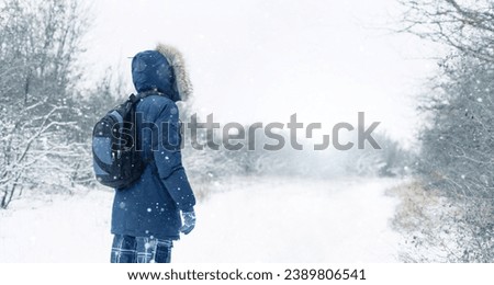 Back view of traveler in a blue jacket with a fur hood and a backpack on the background of a winter landscape during snowfall