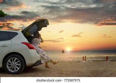 Back view of traveler young woman taking a photo of beautiful sunset while sitting on hatchback car against the copy space sea, clouds and sky background