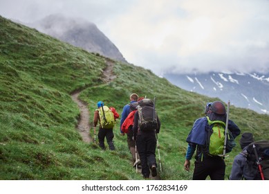 Back view of travelers with backpacks using trekking poles while climbing the grassy hill. Group of male hikers walking on path and heading to foggy mountain. Concept of hiking, climbing and alpinism. - Powered by Shutterstock