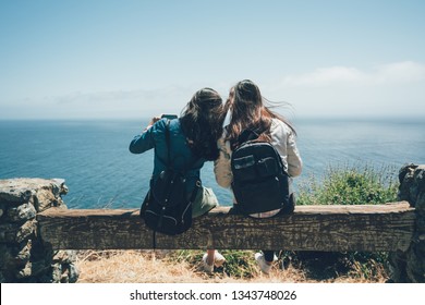 Back View Two Backpackers Friends Sitting On Wooden Bench Chair Railing On Top Of Mountain Using Cellphone Camera App Taking Selfie Picture Outdoors While Travel Hiking In Big Sur Relax Seeing Ocean.