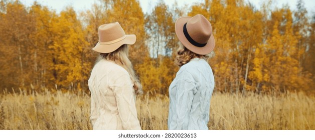Back view of two beautiful young women friends standing together in autumn park, stylish girlfriends in hat and sweater outdoors looking at trees with yellow leaves, nature background