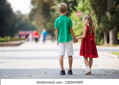 Back view of two cute young blond children, girl and boy, brother and sister walking holding hands on blurred bright sunny park alley green trees bokeh background. Loving siblings relations concept.