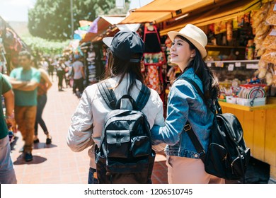 Back View Of Two Female Tourists Visiting The Traditional Market. Beautiful Lady Looking At The Vendor Which They Walked By. Friendship Concept With Young People Shopping In The Holiday Market In LA.