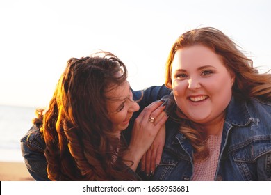 Back view of two freinds , plus size with  thin  girls walking on the fall  beach. Fat woman with strong friend  laughting and holding camera to do selfie. Overweight woman dressed jeans jacket 