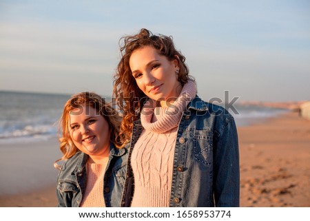 Back view of two freinds , plus size with  thin  girls walking on the spring beach. Fat woman with strong friend  laughting. Overweight woman dressed jeans jacket and pink sweater.