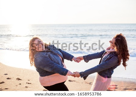 Back view of two freinds , plus size with  thin  girls walking on the spring beach. Fat woman with strong friend  laughting. Overweight woman dressed jeans jacket and pink sweater.