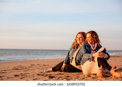Back view two friends , plus size with  thin  girls walking on the spring beach. Fat woman with strong friend  laughting. Overweight woman dressed jeans jacket and pink sweater.