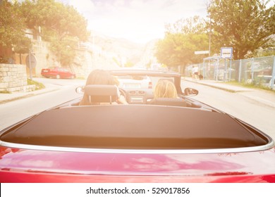 Back View Of Two Girls Driving Red Convertible Car. Sun Effect Applied.