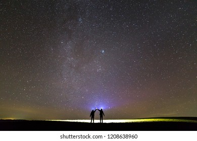 Back view of two men with head flashlights holding raised arms on green grassy field under beautiful dark blue summer starry sky. Night photography, beauty of nature concept, copy space background.