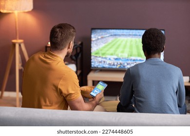 Back view of two men watching football match on TV and holding smartphone with online bets