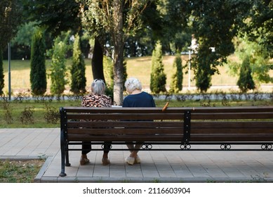 Back View Of Two Old Caucasian Women Girlfriends Relaxing In Park On Summer Day Sitting On Bench Outdoors. Pensioners, Elderly People Leisure In Nature.