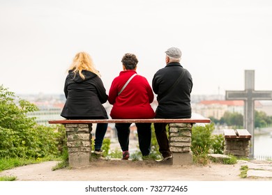 Back View Of Two Overweight Woman And One Older Man Sitting On A Bench Looking Out Over Budapest Hungary. 