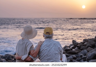 Back view of two romantic seniors embrace at the sea at sunset light sitting on the rocky beach - old elderly couple outdoors enjoying vacations - Powered by Shutterstock