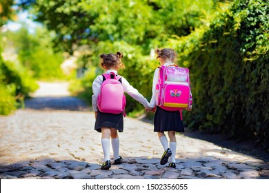 Back View Of A Two School Girls Wearing Backpack Outside The Primary School. Schoolgirl, Elementary School Student Going From School, Graduation, Summer Holidays.