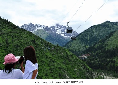 Back view of two tourists looking out over a green mountain valley with cable car gondolas moving toward rocky snow-capped peaks under an overcast sky.