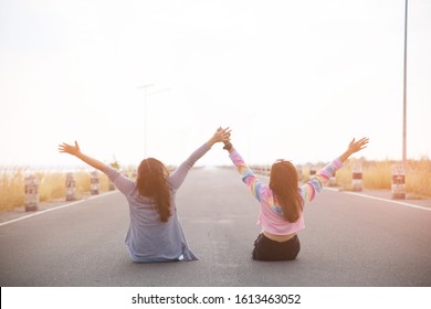 The Back View Of Two Young Woman Sitting On The Road, Looking Far Away And Hands Up.