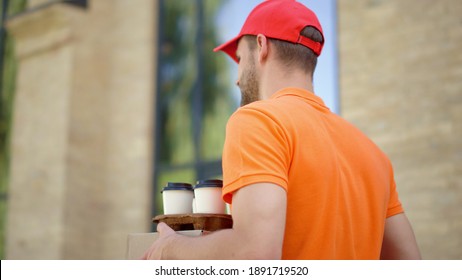 Back View Uniform Courier Going With Box And Coffee Cup Holder Along Street. Delivery Man Looking Around In Urban Background. Cap Guy Working For Delivery Food Service Outdoors.