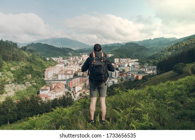 Back view of unknown man looking the valley view. He is wearing a backpack and dark sportswear. Village in a valley rounded by mountains and blue sky in background