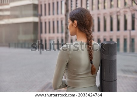 Back view of unrcognizable fitness model dressed in activewear has combed pigtail carries rolled karemat looks around takes break after cardio training surrounded by cityscrapers blurred background