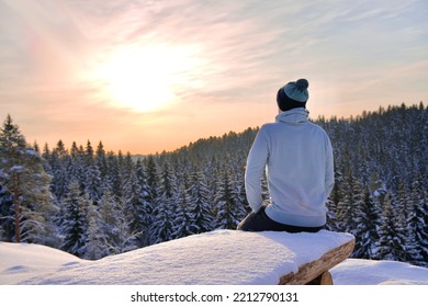 back view of unrecognisable person, man in hat at snowy winter landscape, forest trees in snow sitting on bench at sunny cold frosty weather Sunrise in mountain.