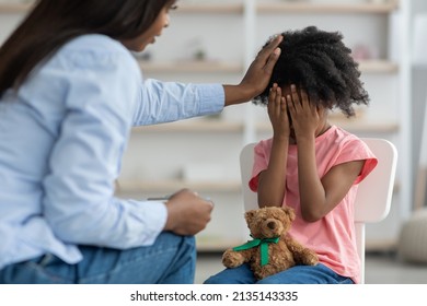 Back view of unrecognizable african american woman comforting emotional curly girl preschooler crying during therapy session with child psychologist, carrying teddy bear on her lap, clinic interior