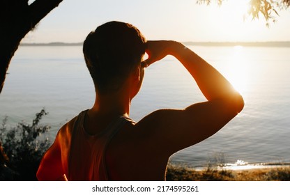 Back View Of Unrecognizable Athletic Man Enjoying Beautiful Sunset At Lake. Inspirational Guy Looking At Setting Bright Sun Outdoors On Sunny Evening.