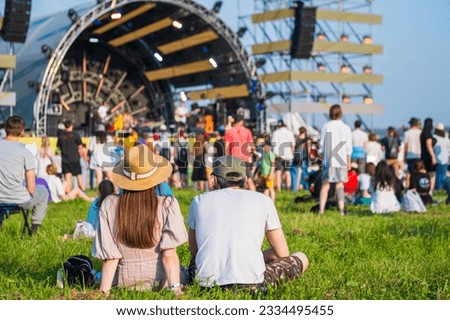 Back view of unrecognizable couple in casual clothes sitting on ground in front of crowd and stage during music festival on sunny summer day