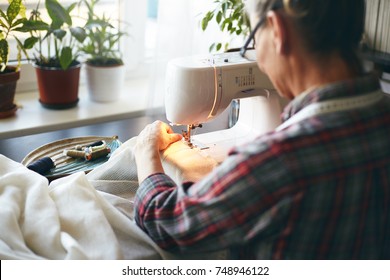 Back View Of Unrecognizable Elderly Needlewoman In Plaid Shirt Sewing Kitchen Curtains Using Stitching Machine. Senior Female Dressmaker Spending Early Morning, Repairing Or Altering Clothing Item