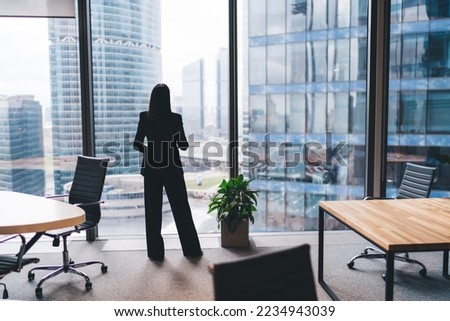Back view of unrecognizable female entrepreneur in formal clothes standing near glass wall and office furniture at workplace while looking away at cityscape