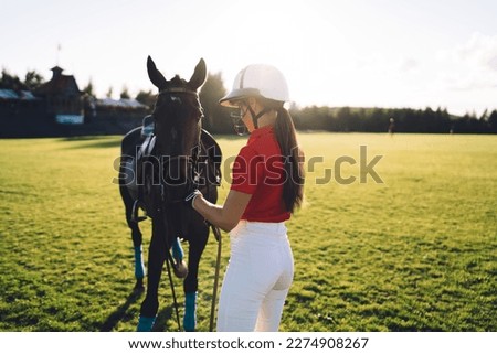 Back view of unrecognizable female equestrian in helmet and uniform standing near chestnut horse during training on green meadow against cloudless sky