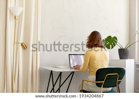 Back view of unrecognizable female in smart casual clothes sitting at desk and browsing data on netbook with empty screen while working in stylish workplace at home