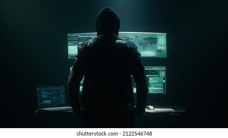 Back View Of Unrecognizable Man In Hoodie Standing Near Desk And Reading Stolen Data From Computer, Monitors In Dark Room Before Massive Cyber Attack On Servers.