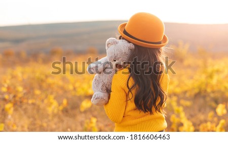 Back view of unrecognizable sad little girl in yellow sweater and hat cuddling teddy bear while standing alone in blooming summer field