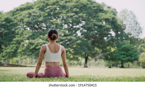 Back view of an unrecognizable slender Asian young woman sitting on the grass in the lotus position and raising hands up outside in city park with the big trees background. 