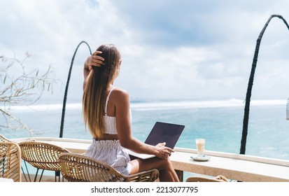 Back View Of Unrecognizable Slim Female Freelancer Sitting At Table And Touching Hair While Working Online Via Laptop On Terrace Near Endless Blue Ocean In Bali